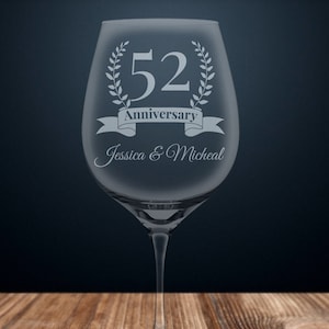 May include: A clear wine glass with etched text. The text reads "52 Anniversary" with a laurel wreath and banner. Below the banner is "Jessica & Micheal". The glass is on a wooden surface with a dark blue background.