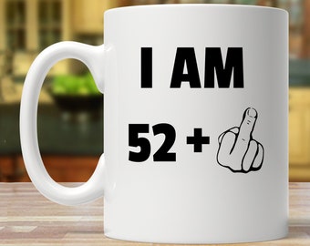 以下が含まれることがあります： 白いセラミック製のコーヒーカップで、黒いグラフィックが「I AM 52+」と中指の絵文字で書かれています。