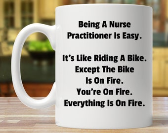 May include: White ceramic mug with black text that reads "Being A Nurse Practitioner Is Easy. It's Like Riding A Bike. Except The Bike Is On Fire. You're On Fire. Everything Is On Fire."