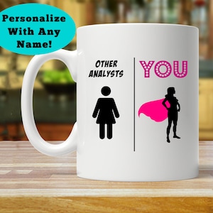 May include: White ceramic coffee mug with a black and white illustration of a woman with a cape. The text "Other Analysts" is on the left side of the mug and "YOU" is on the right side of the mug.