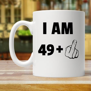 May include: White ceramic coffee mug with black text that reads "I AM 49+ " and a black and white illustration of a middle finger.