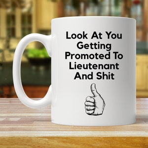 May include: White ceramic coffee mug with black text that reads "Look At You Getting Promoted To Lieutenant And Shit" with a thumbs up icon.