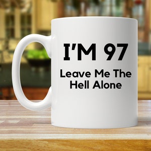 Peut inclure: Mug en céramique blanche avec une anse noire. Le mug porte l'inscription "I'M 97 Leave Me The Hell Alone" en lettres noires grasses. Le mug est posé sur une surface en bois.