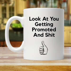May include: White ceramic coffee mug with a black graphic that says "Look At You Getting Promoted And Shit" with a thumbs up icon.