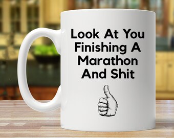 May include: White ceramic coffee mug with a black graphic that reads "Look At You Finishing A Marathon And Shit" with a thumbs up icon.