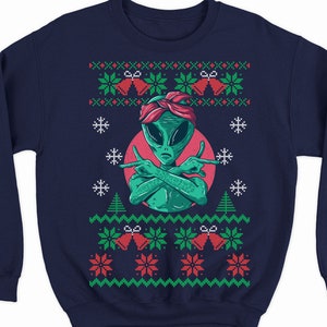May include: A navy blue sweatshirt with a green alien wearing a red bandana and making a peace sign with both hands. The alien is in the center of a red circle with a white outline. The sweatshirt has a red and green Christmas pattern with snowflakes and bells.