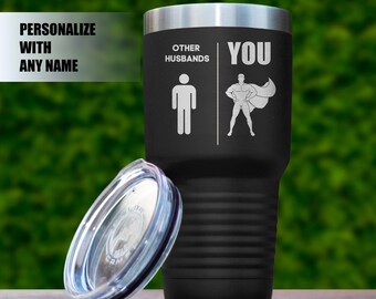May include: Black stainless steel tumbler with a lid. The tumbler has the text "Other Husbands" and a stick figure on the left side. The right side has the text "You" and a stick figure wearing a cape.