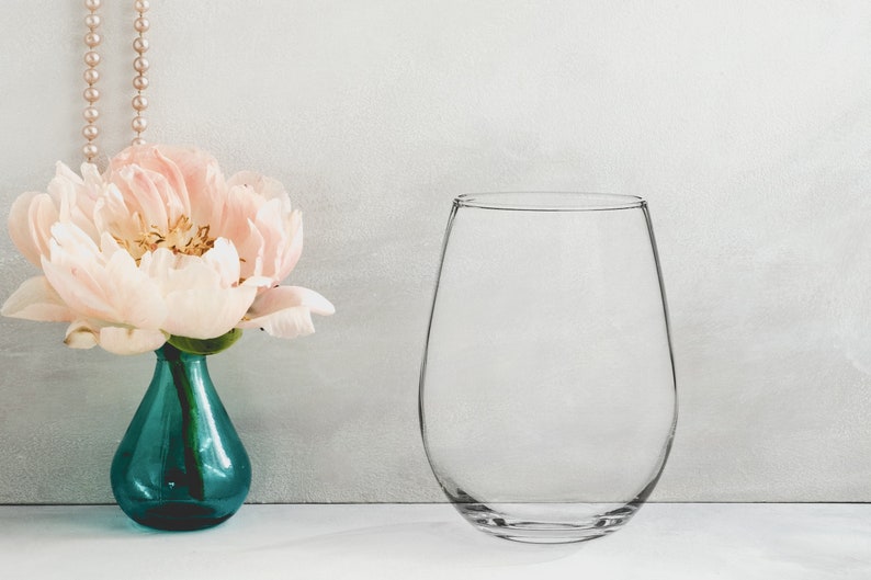 May include: A clear glass stemless wine glass sits on a white surface next to a light pink peony in a teal blue vase.