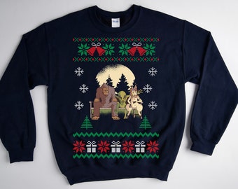 May include: Navy blue sweatshirt with a whimsical Christmas design. The graphic features Bigfoot, an alien, and a deer-like creature sitting on a bench in front of a full moon. The top and bottom of the sweatshirt have a festive pattern with bells, snowflakes, and gift boxes.