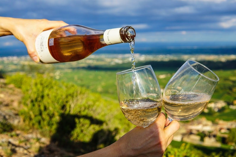May include: Two glasses of white wine being poured from a bottle of rose wine. The glasses are held by hands and the background is a scenic view of a vineyard.