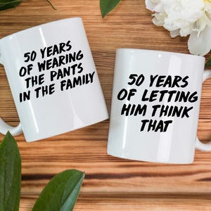 May include: Two white coffee mugs with black text. The mug on the left says "50 Years of Wearing the Pants in the Family". The mug on the right says "50 Years of Letting Him Think That".