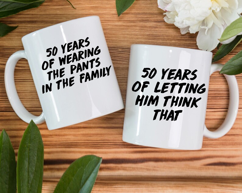 May include: Two white coffee mugs with black text. The mug on the left says "50 Years of Wearing the Pants in the Family". The mug on the right says "50 Years of Letting Him Think That".
