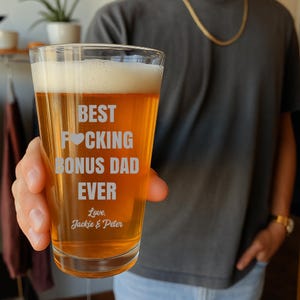 以下が含まれることがあります： ビールと泡が入った透明なパイントグラスを人が持っています。「BEST F*CKING BONUS DAD EVER」と「Love, Jackie & Peter」の文字が刻まれています。