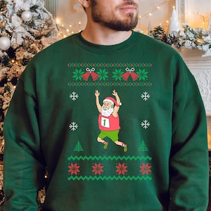 May include: A dark green Christmas-themed sweatshirt featuring a running Santa Claus graphic. The design includes snowflakes, Christmas bells, and a red and green pattern. The Santa Claus is wearing a red hat and vest with the number 1.