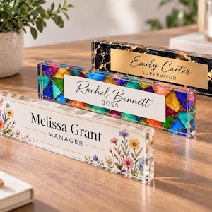 May include: Three rectangular acrylic desk nameplates. The front one features a floral print with the text Melissa Grant MANAGER. The middle one has a colorful stained-glass pattern with the text Rachel Bennett BOSS. The back one has a black and gold marble design with the text Emily Carter SUPERVISOR.