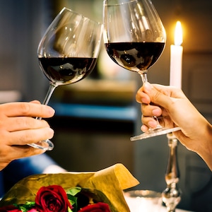 May include: Two glasses of red wine are raised in a toast, with a bouquet of red roses in the foreground. The glasses are clear and have a long stem. The wine is a deep red color. The roses are arranged in a brown paper wrapper.