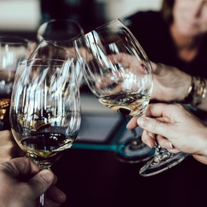 May include: Four clear glass wine glasses filled with white wine, being raised in a toast. The glasses are held by multiple hands.