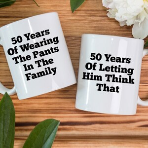 May include: Two white ceramic mugs with black text. One mug reads "50 Years Of Wearing The Pants In The Family." The other mug says "50 Years Of Letting Him Think That."