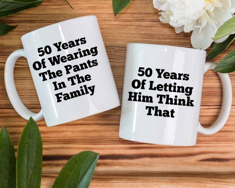 May include: Two white ceramic mugs with black text. One mug reads "50 Years Of Wearing The Pants In The Family." The other mug says "50 Years Of Letting Him Think That."