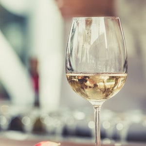 May include: A glass of white wine in a clear glass with a stem. The wine is a pale yellow color and the glass is partially filled.