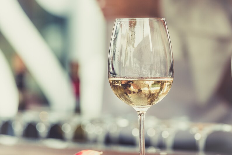 May include: A glass of white wine in a clear glass with a stem. The wine is a pale yellow color and the glass is partially filled.