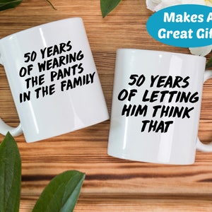 May include: Two white coffee mugs with black text. The mug on the left says "50 Years of Wearing the Pants in the Family". The mug on the right says "50 Years of Letting Him Think That".