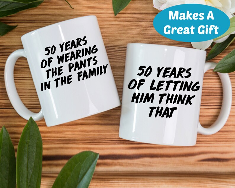 May include: Two white coffee mugs with black text. The mug on the left says "50 Years of Wearing the Pants in the Family". The mug on the right says "50 Years of Letting Him Think That".