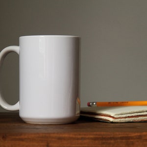 Op de afbeelding: Een witte keramische mok met een handvat, die op een houten tafel staat naast een stapel notitieboekjes en een geel potlood.