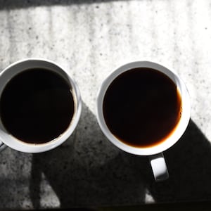 Op de afbeelding: Twee witte keramische mokken gevuld met zwarte koffie, op een lichtgrijs aanrecht.