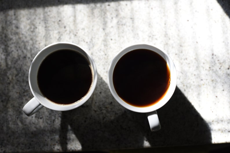 Op de afbeelding: Twee witte keramische mokken gevuld met zwarte koffie, op een lichtgrijs aanrecht.