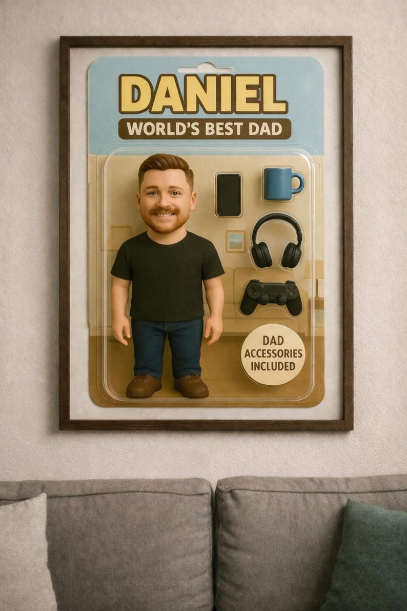 May include: A framed novelty toy package featuring a figurine of a man in a black t-shirt and jeans. The package includes miniature accessories: a phone, mug, headphones, and game controller. Text reads "DANIEL WORLD'S BEST DAD."
