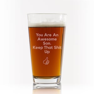 May include: A clear glass pint glass with a brown liquid inside. The glass has the text "You Are An Awesome Son. Keep That Shit Up" and a thumbs up icon etched on the side.