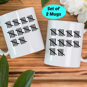 May include: Set of two white ceramic mugs with black tally marks. The mugs are on a wooden surface. The text "Set of 2 Mugs" is on a blue circle.