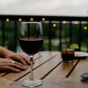 May include: A glass of red wine sits on a wooden table with a blurry background of a patio with string lights.