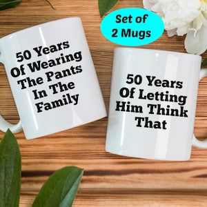 May include: Two white ceramic mugs with black text. One mug reads "50 Years Of Wearing The Pants In The Family." The other mug reads "50 Years Of Letting Him Think That." A turquoise oval with "Set of 2 Mugs" is in the background.