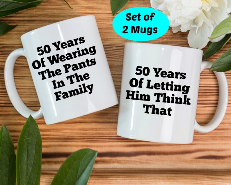 May include: Two white ceramic mugs with black text. One mug reads "50 Years Of Wearing The Pants In The Family." The other mug reads "50 Years Of Letting Him Think That." A turquoise oval with "Set of 2 Mugs" is in the background.