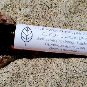 Peut inclure: Un petit flacon en verre brun avec un bouchon noir et une étiquette blanche. L'étiquette présente un motif de feuille et le texte "Hollywood Hippie Juice C.T.F.D. - Calming Blend Basil, Lavender, Orange, Patchouli & Peppermint essential oils @thehollyw.podhippie".