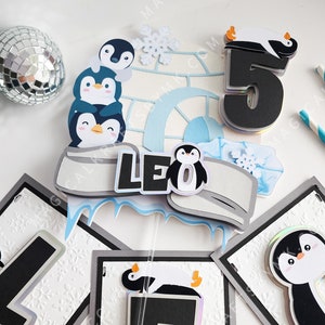 May include: A penguin-themed cake topper with a blue and white igloo, penguins, and the name "Leo" in black letters. The topper is surrounded by a banner with the letters "L", "E", and "O" in black and iridescent letters.