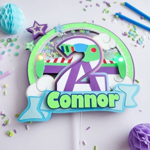 May include: A purple and green cake topper featuring a number 2, a green alien, and the name "Connor" on a blue banner. The topper is decorated with glitter and sequins.