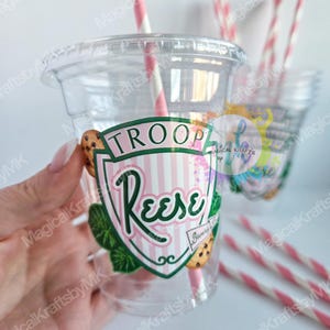 May include: Clear plastic cup with a lid and a pink and white striped straw. The cup has a label that says "TROOP Reese" with a cookie graphic. Additional cups and straws are visible in the background.