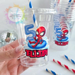May include: A clear plastic party cup featuring a graphic of a red and blue superhero character sitting on a blue inner tube, a large blue number five, and the name Elijah in red block letters. A blue and white striped paper straw is inside.