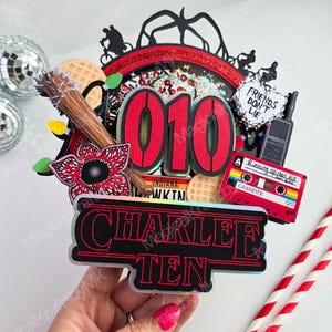 May include: A multi-layered cake topper with a red and black color scheme. It features the number "010", the name "CHARLEE TEN", a Demogorgon, a cassette tape, and the text "FRIENDS DON'T LIE".