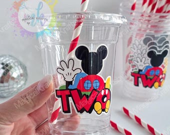 May include: A clear plastic cup featuring a colorful decal of a cartoon mouse silhouette, red shorts with blue buttons, and the word TWO in bold black letters. A red and white striped paper straw is inserted into the lid of the cup.
