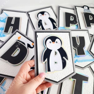 May include: A penguin-themed birthday banner with the word "Happy" spelled out in black letters. Each letter is on a separate white card with a penguin illustration and a gray border. The cards are connected with string.