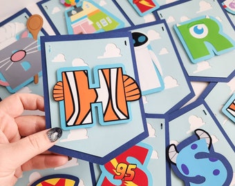 Könnte beinhalten: Eine Sammlung bunter Bannerstücke mit Zeichentrickfiguren und Buchstaben. Der Buchstabe "H" ist orange und weiß und ähnelt einem Clownfisch. Andere Banner zeigen Buchstaben, Zahlen und Figuren aus Animationsfilmen vor blauem Himmel.