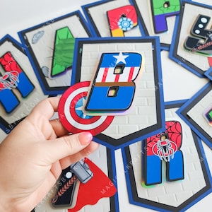 May include: Superhero-themed banner with letters and character emblems. The letter "B" features Captain America's colors and a shield. Other letters include Spider-Man, Thor, and Iron Man. The banner has a blue border and a brick-like background.