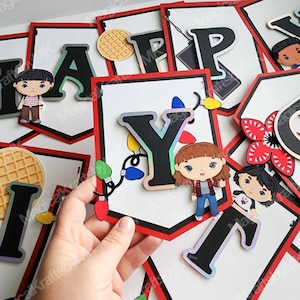 May include: A banner with black letters and red trim, featuring characters and symbols. The letters spell out "HAPPY BIRTHDAY". The banner includes character cutouts and decorative lights.