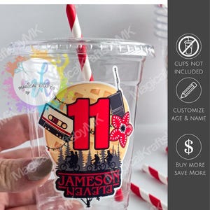 May include: A clear plastic cup featuring a large sticker with the number 11 in red, a cassette tape, a walkie-talkie, a red flower, and a silhouette of three people on bicycles in a forest. The sticker includes the name Jameson and the word Eleven.