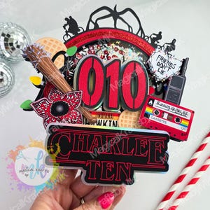 May include: A colorful cake topper with a Stranger Things theme. It features the number "010" in red, a Demogorgon, a cassette tape, and the text "CHARLEE TEN". The topper includes a waffle and the phrase "FRIENDS DON'T LIE".