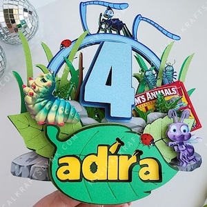 May include: A colorful, layered paper cake topper featuring a blue number 4, cartoon insects, and the name "adira" in yellow. The design includes a caterpillar, ladybugs, and a book titled "M's Animals".